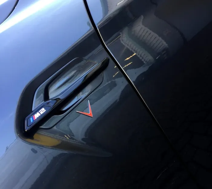 A car door contains a black handle and an emblem displaying "M2" with colored stripes, set against a shiny, dark-colored exterior, reflecting nearby buildings.