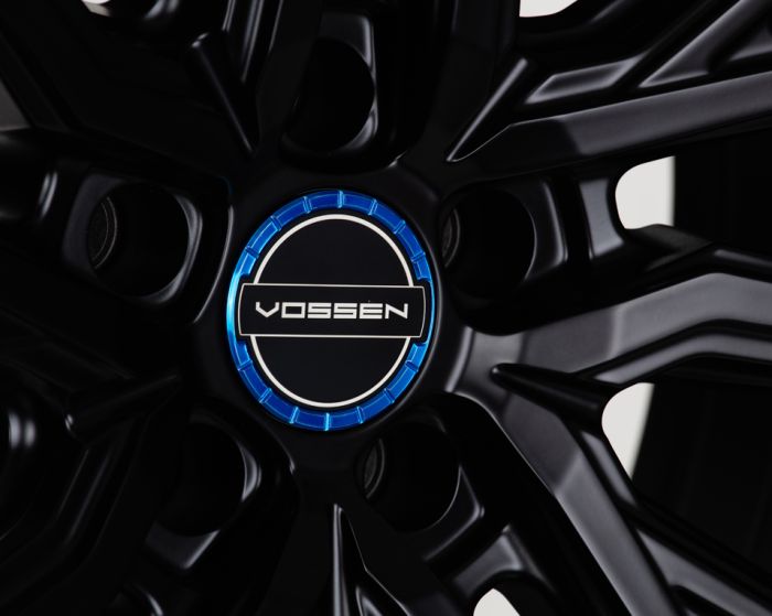 Black alloy wheel hub with the text "VOSSEN" on a central circular emblem, bordered by a blue ring. The wheel features a sleek, angular design in a close-up view.