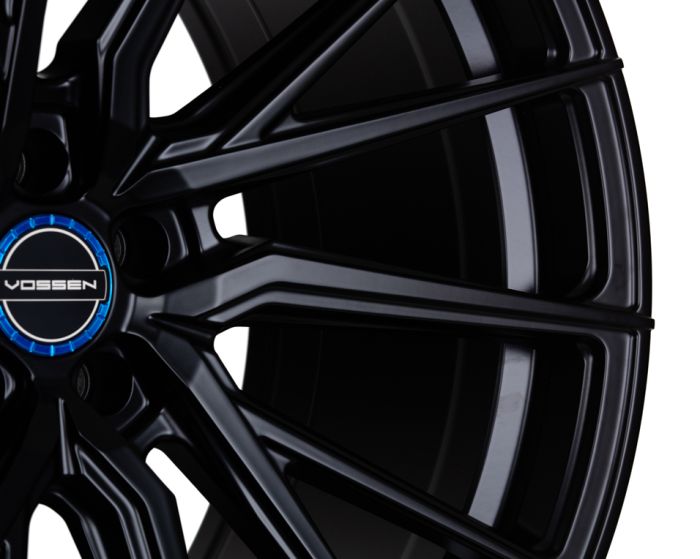 A black alloy wheel with intricate spokes displays the "VOSSEN" logo in the center, highlighted by a blue accent, against a plain, bright background.