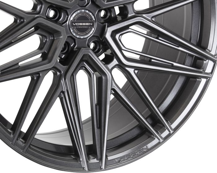 Alloy wheel with intricate, angular spoke design; center cap reads "VOSSEN HYBRID FORGED." Displayed in a studio setting, showcasing the glossy metallic finish and craftsmanship.