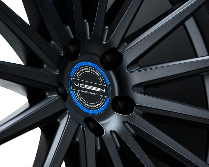 A metallic alloy wheel with long spokes is displayed, featuring a central blue and black emblem labeled “VOSSEN HYBRID-FORGED” with text "VOSSEN WHEELS EST 2006 U.S.A. PRECISION - PERFORMANCE - PRECISION" in a clean, close-up view.