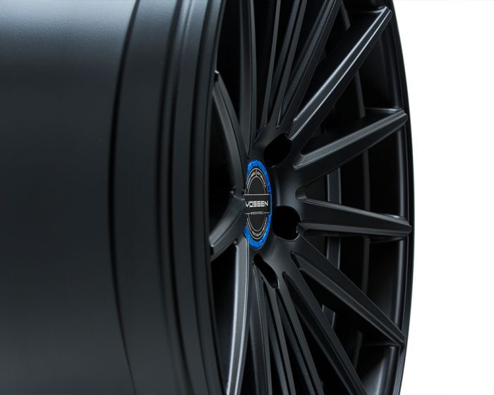 A matte black alloy wheel, featuring multiple thin spokes converging at a metallic blue center cap labeled "VOSSEN FORGED," is displayed against a plain white background.