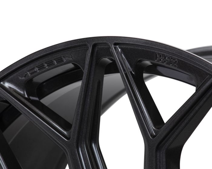 Black metallic wheel rim features triangular spokes, labeled "VOSSEN" and "HF-2," displayed in close-up view, highlighting modern design elements and smooth finish.