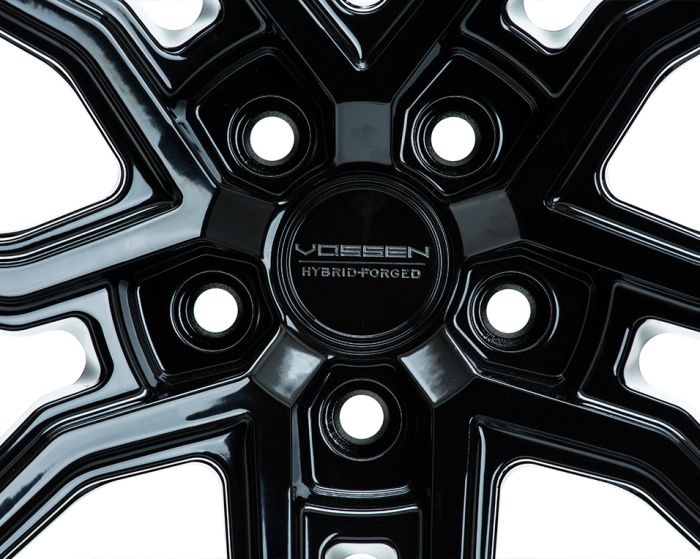 Glossy black wheel rim features five bolt holes and intricate design; central hub reads "VOSSEN HYBRID FORGED"; close-up shot highlights reflective surface and geometric patterns.