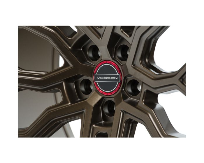 Bronze-colored alloy wheel with intricate spokes, featuring a center cap labeled "VOSSEN" in a red and black circle. The setting is a close-up view, emphasizing design details.