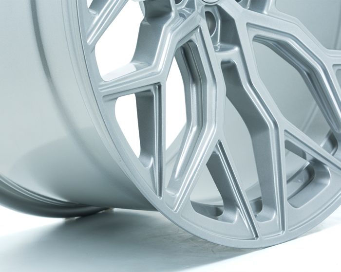 A silver alloy wheel with intricate, angular spoke design rests on a smooth white surface, highlighting its modern and sleek aesthetic.