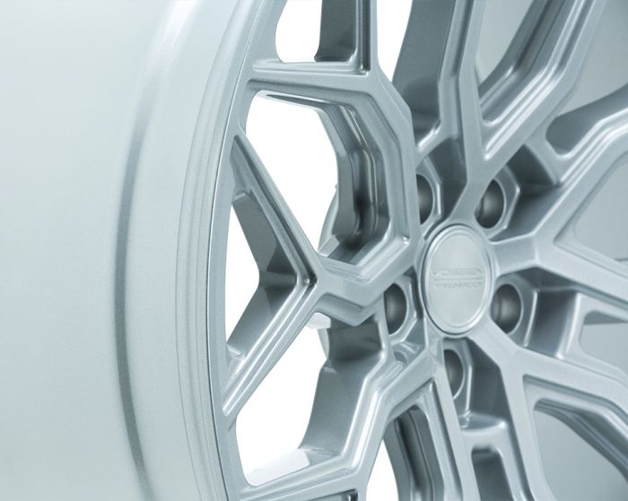 A silver alloy wheel with a geometric spoke design rests stationary, highlighting precision engineering. The center cap features the text: "VOSSEN HYBRID FORGED." The background is plain, emphasizing the wheel's details.