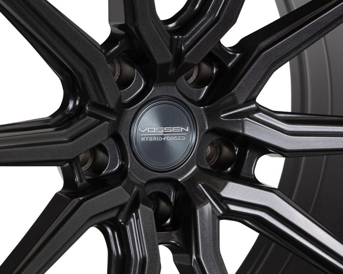 A black, detailed car wheel rim with a central cap labeled "VOSSEN HYBRID FORGED," featuring a multi-spoke design, is shown in a close-up view.