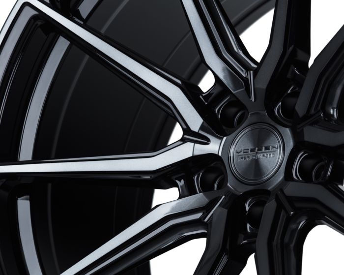 A glossy black alloy wheel features intricate spoke design and the text "Vossen Hybrid Forged" at the center, amidst a shadowed background.