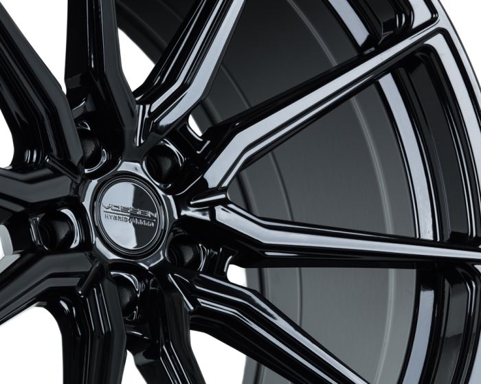 A glossy black alloy wheel with intricate spokes, displaying the text "VOSSEN HYBRID FORGED" on its central cap, against a minimalist background.