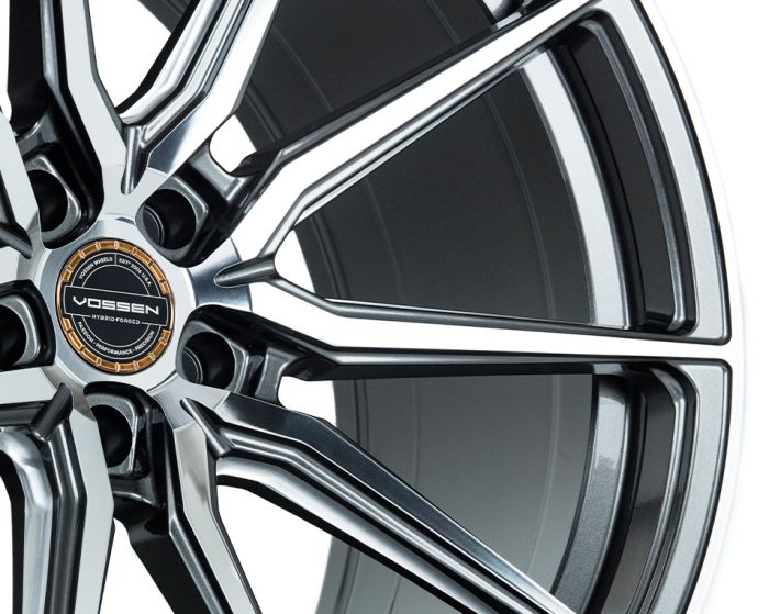 A silver alloy wheel with intricate, modern spoke design, displaying the text "Vossen Hybrid Forged" on a detailed center cap in a studio setting.