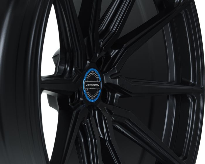 A black metal car wheel is shown with a multi-spoke design in a studio setting. The center cap features text: "VOSSEN," "HYBRID FORGED," and "FLOW FORM TECHNOLOGY."