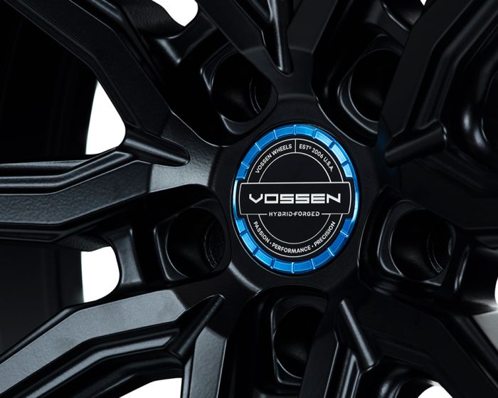 A black alloy wheel with "VOSSEN" branding on a blue center cap, surrounded by five spokes. Text includes "VOSSEN WHEELS," "EST 2006 USA," "HYBRID-FORGED," and "PASSION • PERFORMANCE • PRECISION."