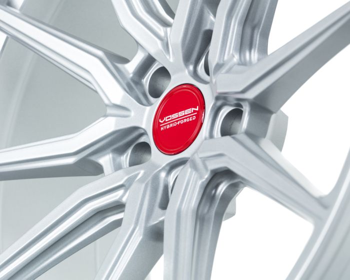 Silver alloy wheel featuring a multi-spoke design, with a red center cap displaying the text "Vossen Hybrid Forged," set against a plain, light background.