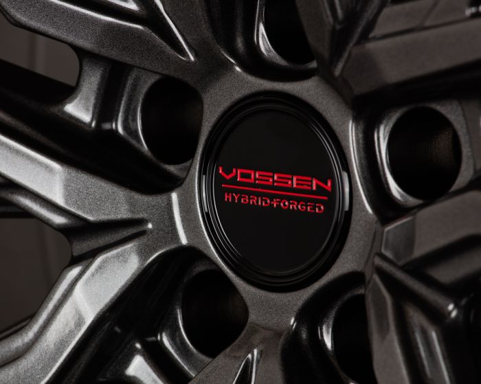 A metallic wheel rim displays the red text "VOSSEN HYBRID FORGED" on its black center cap, set against a shiny, contoured surface with dark grooves.