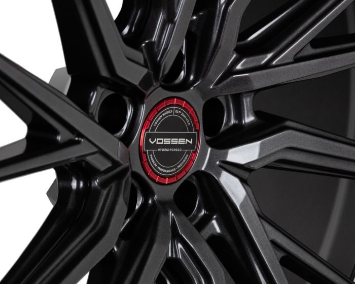 A black alloy wheel with intricate spokes, featuring a red center cap labeled "VOSSEN HYBRID-FORGED" surrounded by text: "VOSSEN WHEELS EST. 2006, USA," "PASSION," "PERFORMANCE," "PRECISION."