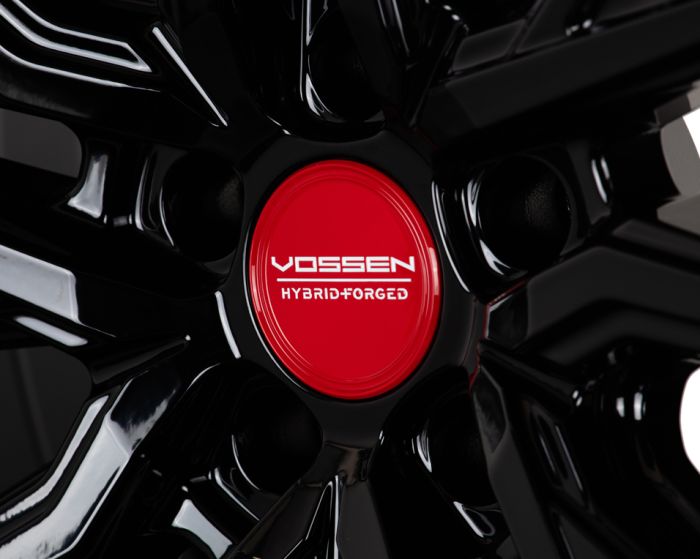 A black wheel rim with a glossy finish features a red center cap labeled "VOSSEN HYBRID FORGED," set against a dark background.