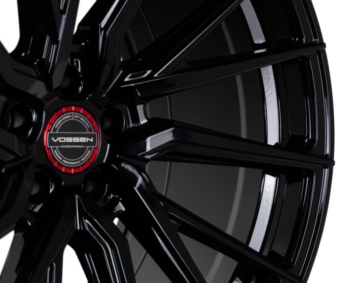 A glossy black alloy wheel with angular spokes, featuring a red center cap with "VOSSEN HYBRID FORGED" text, set against a plain white background.