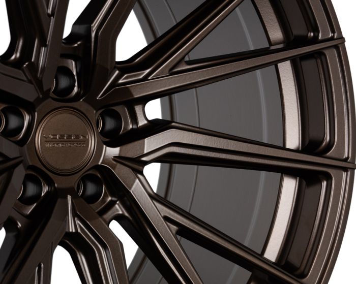 Bronze alloy wheel with intricate spoke design, displaying the logo "Vossen Hybrid Forged" at the center; set against a minimalistic, light background.