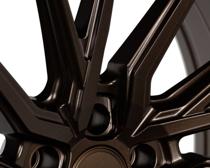 A bronze-colored alloy wheel displays a close-up of its intricate, angular spoke design, highlighting the metallic finish. The focus is on the geometric patterns and smooth texture.