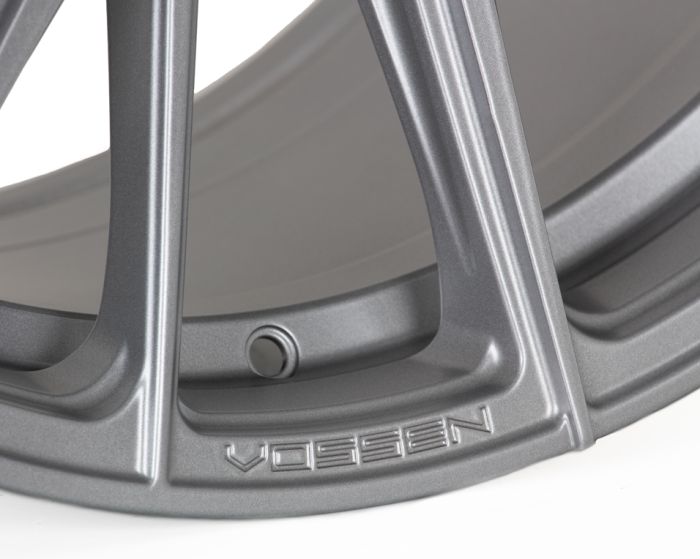 A metallic car wheel rim with prominent spokes; the brand name "VOSSEN" is embossed near the edge, highlighting its design. The background is neutral, showcasing its details.