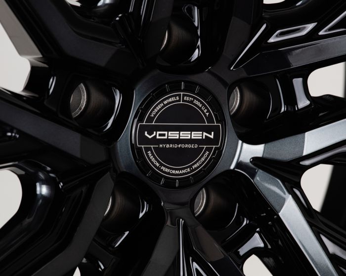 A black alloy wheel with a glossy finish, featuring Vossen branding at the center cap, surrounded by angular spokes. Text reads: "VOSSEN WHEELS EST'D 2006 U.S.A. HYBRID FORGED PASSION • PERFORMANCE • PRECISION."