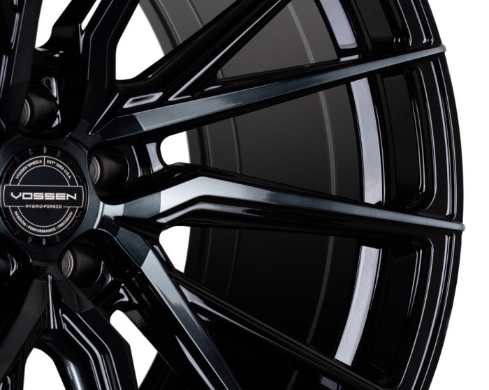 A sleek black alloy wheel with intricate spokes displays the logo "Vossen Hybrid Forged" and the text "Vossen Wheels Est 2006 USA" in a close-up view, highlighting its glossy finish.