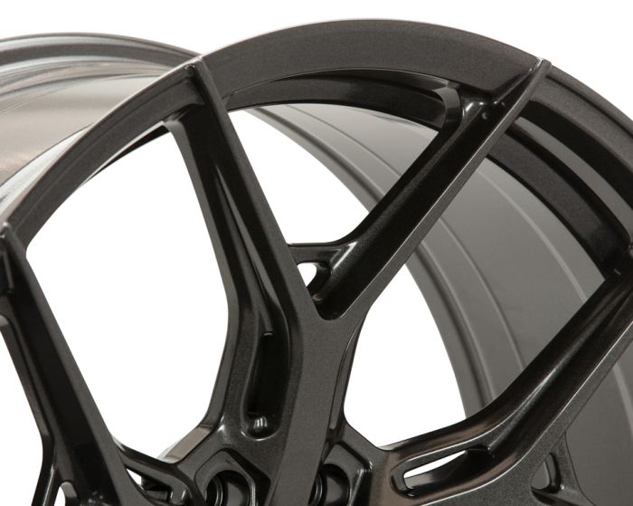 A close-up of a metallic, dark-colored car wheel rim with intricate, angular spoke design. The background is a neutral, light shade, highlighting the rim's sleek contours.