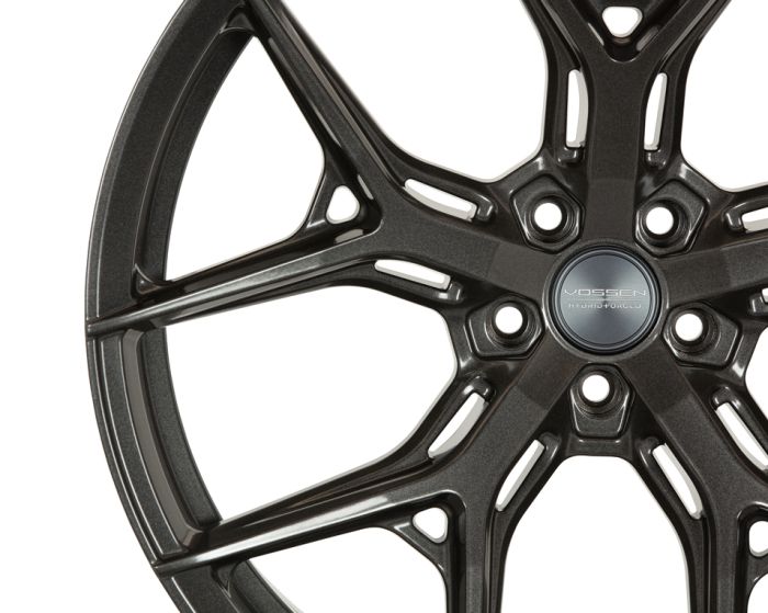 A metallic car wheel rim with intricate, angular spokes features the text "VOSSEN HYBRID FORGED" at its center, set against a plain white background.