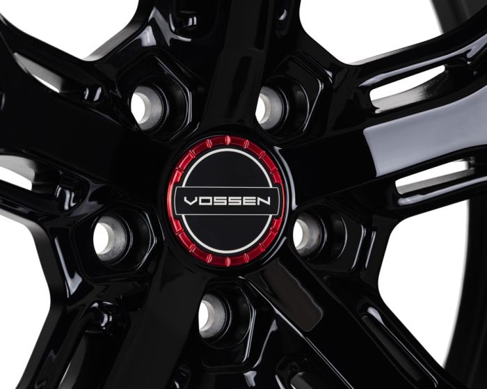 A glossy black alloy wheel features the logo "VOSSEN" at its center, bordered by a red ring, within a studio-lit environment.