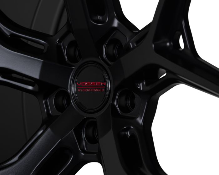 A black wheel rim, labeled "VOSSEN HYBRID FORGED," features sleek, intersecting spokes in a high-contrast environment, emphasizing its glossy finish and modern design.