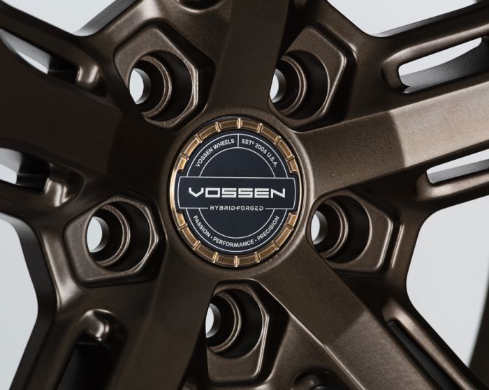 Bronze alloy wheel showcasing intricate design with central logo. Text reads: “VOSSEN WHEELS EST. 2006 U.S.A. VOSSEN HYBRID FORGED PASSION - PERFORMANCE - PRECISION.” Viewed against a neutral background.