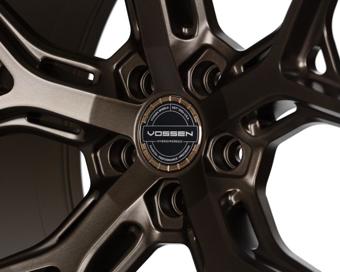 A bronze alloy wheel with a five-spoke design displays the text: "VOSSEN WHEELS EST 2006 U.S.A., VOSSEN, HYBRID-FORGED, PASSION - PERFORMANCE - PRECISION" in a car showroom setting.