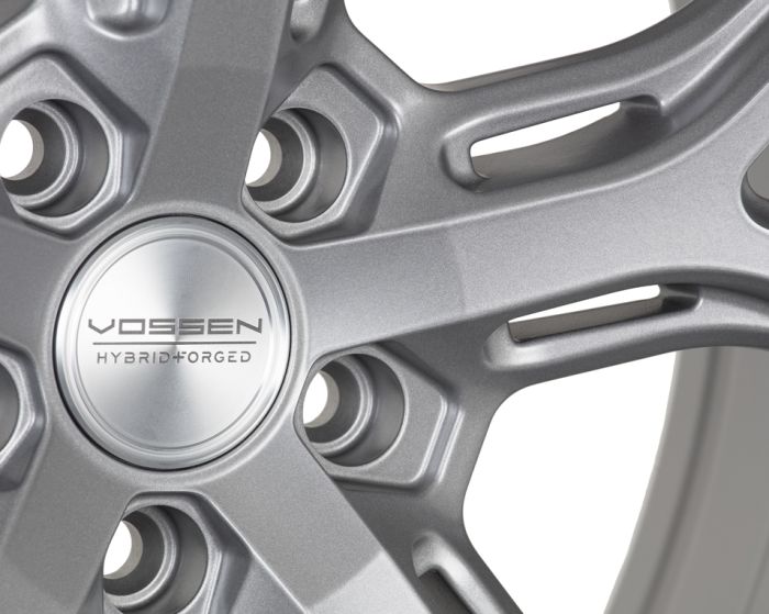 A silver alloy wheel displays a detailed design with angular spokes, featuring a central cap labeled "VOSSEN HYBRID FORGED" in a close-up view, highlighting craftsmanship.