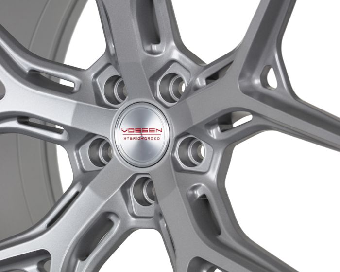 A metallic car wheel features intricate silver spokes radiating from a center hubcap displaying the text "VOSSEN HYBRID FORGED" in a bold, red font, set against a white background.