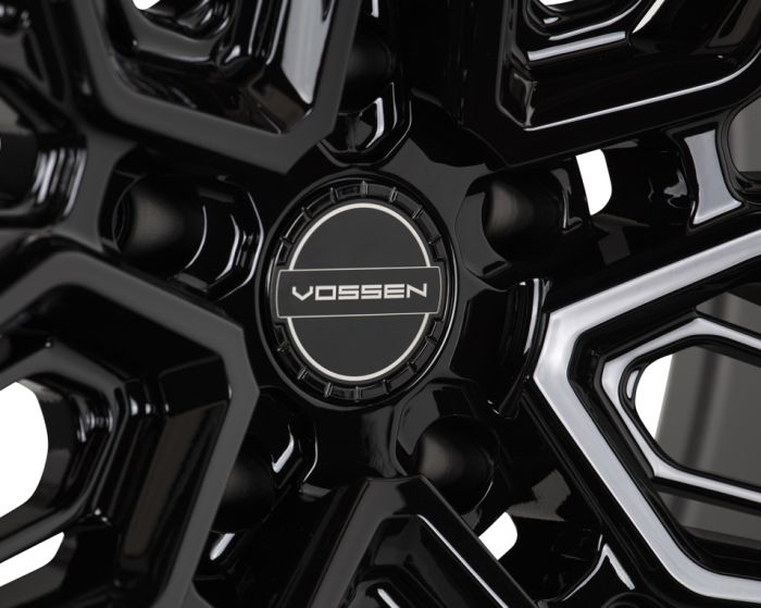 A glossy black wheel with intricate geometric spokes, centered on a round cap labeled "VOSSEN," set against a blurred industrial backdrop.