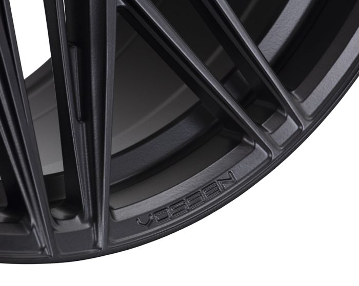 A close-up of a black wheel rim displaying the word "VOSSEN" embossed on the brushed metallic surface, showcasing intricate, angular spokes in a modern, industrial style.