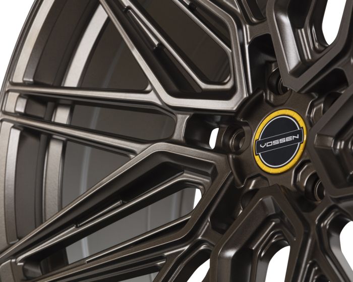 An intricate black alloy wheel features multiple spokes, with a central logo displaying "VOSSEN" against a yellow and black circular background; background is neutral, highlighting the wheel's design.