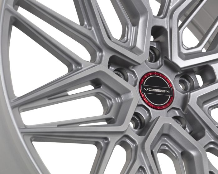 A silver alloy wheel with intricate geometric spokes features a central cap displaying the text "VOSSEN" on a black background, set within a red circular accent.