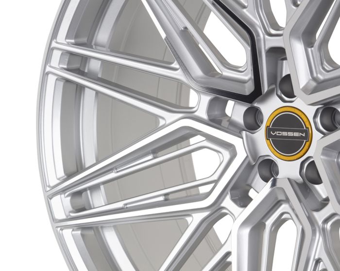 A polished, intricate alloy wheel with a multi-spoke pattern displays the logo "VOSSEN" at its center, surrounded by sleek metallic reflections in a studio setting.