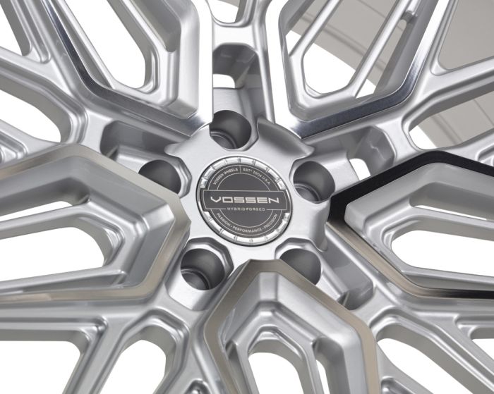 A silver alloy wheel features a complex geometric design; the center cap reads: "Vossen Hybrid Forged, Vossen Wheels, Est 2006, USA, Passion - Performance - Precision." It’s set against a white background.