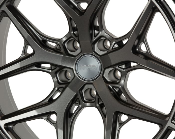 A metallic car wheel rim features intricate, interlacing spokes with a central hubcap. The hubcap displays text: "VOSSEN HYBRID FORGED." The background is a plain, neutral color.