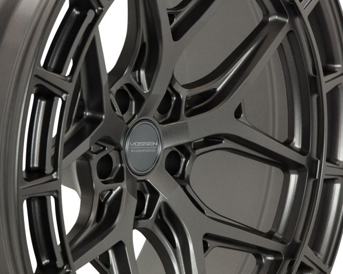 A detailed Vossen hybrid-forged wheel displayed at an angle, showing intricate spoke design, in a studio setting. The wheel has a matte finish with the text "VOSSEN HYBRID FORGED" on the center cap.