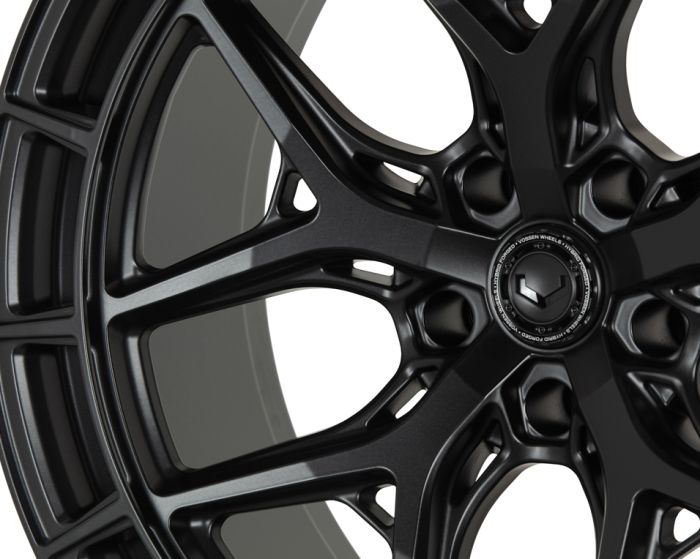 A black alloy wheel with intricate, angular spokes in a close-up view. The text around the center reads "FORGED VOSSEN WHEELS • VOSSEN FORGED • DESIGN". The background is plain white.