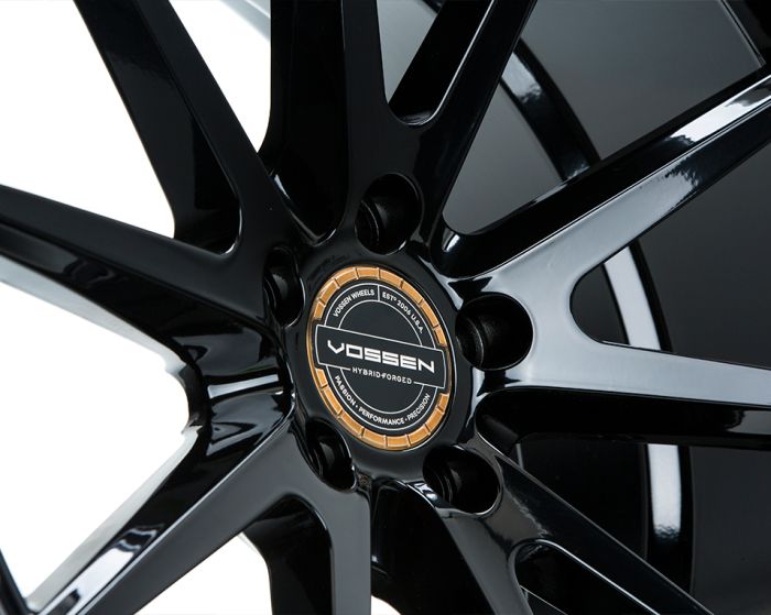 A glossy black alloy wheel with a multi-spoke design shows the Vossen logo and text, "HYBRID-FORGED," "VOSSEN WHEELS EST 2006 USA," "PRECISION - PERFORMANCE - PASSION," in a studio setting.