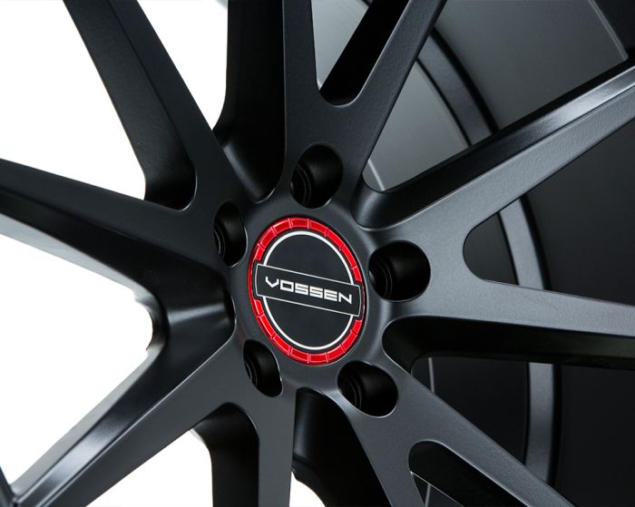 A black alloy wheel with a red center cap displays the logo "VOSSEN," featuring a modern, multi-spoke design in a well-lit setting.