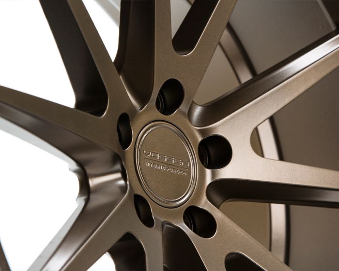 A bronze alloy car wheel displays a sleek, multi-spoke design. The center cap features engraved text reading "VOSSEN" and additional smaller text, within a studio-lit environment.