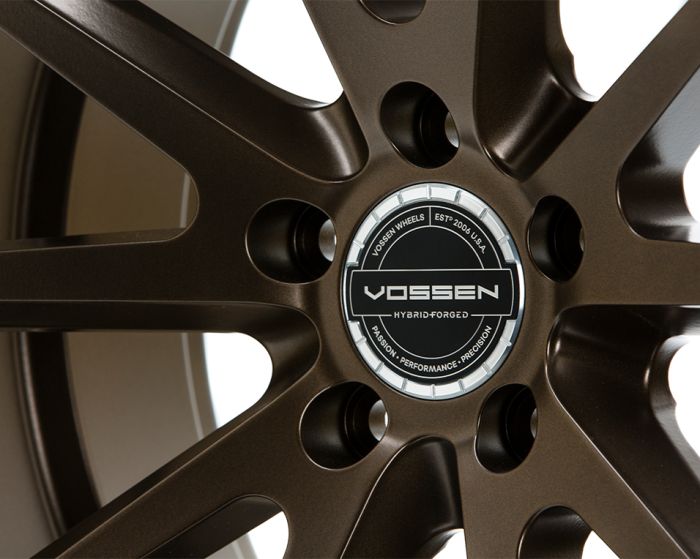 A dark brown metallic wheel rim, embellished with the logo "VOSSEN HYBRID FORGED" centered amidst sleek spokes. Text includes "VOSSEN WHEELS, EST 2006 USA, PASSION - PERFORMANCE - PRECISION."