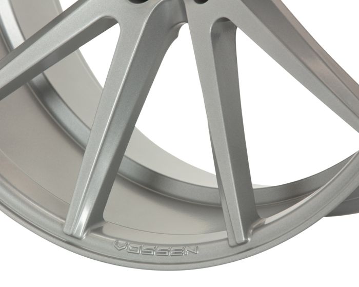 A metallic Vossen car wheel rim with sleek, angular spokes sits against a plain white background. The brand name "VOSSEN" is engraved near the rim edge.