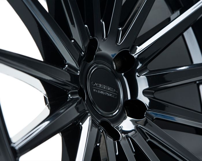 A close-up of a glossy black car wheel with multiple spokes. Center cap displays "VOSSEN" and "HYBRID FORGED" text, highlighting brand and manufacturing details.